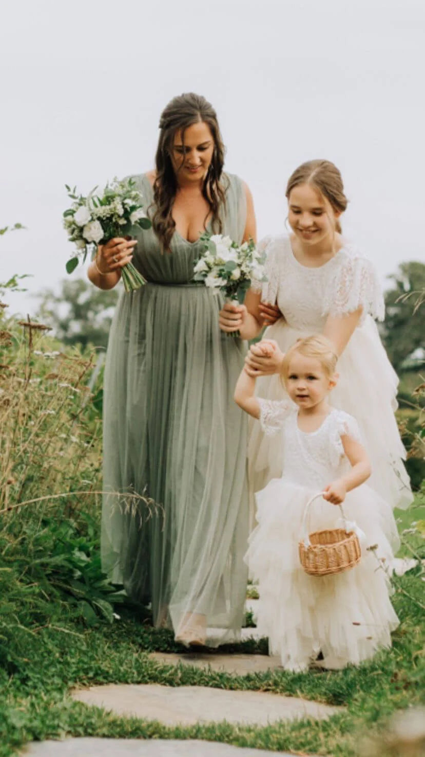 A bride in a white lace wedding dress stands beside a woman in a flowing sage green bridesmaid gown, with a young flower girl in a white dress holding a small basket outdoors on a stone path.