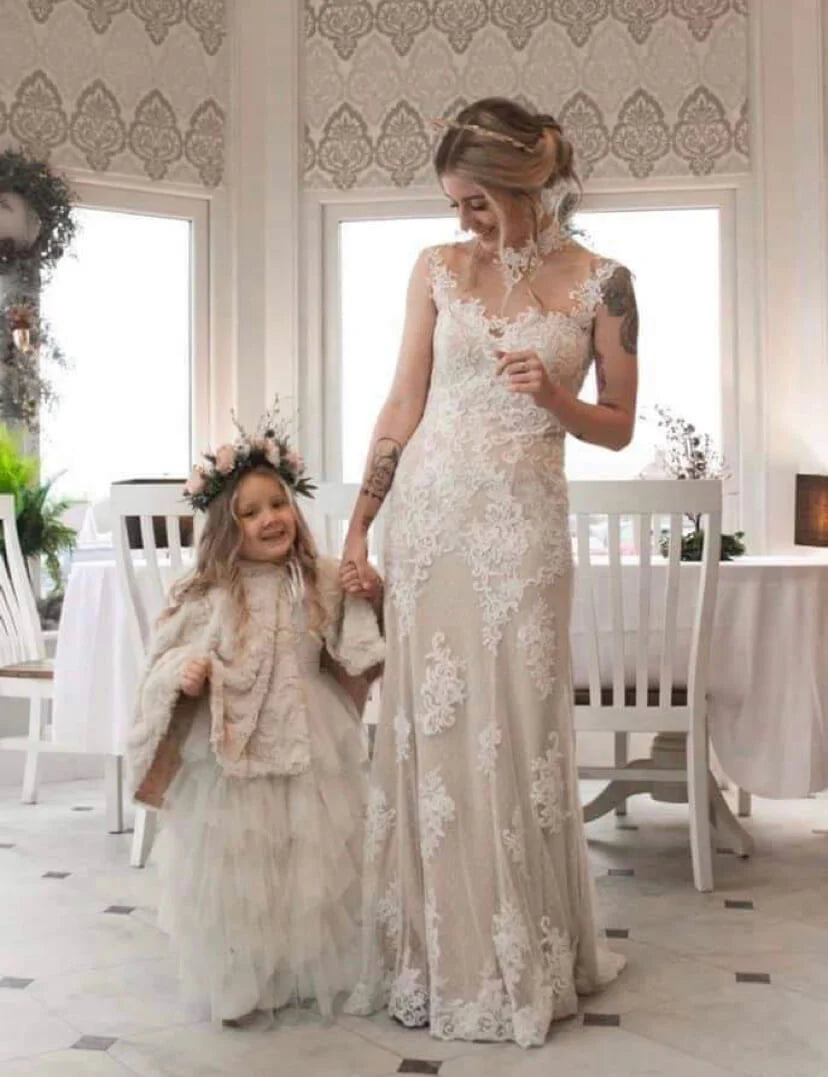 A woman in a sleeveless lace wedding gown holding hands with a young girl wearing a matching tulle dress and floral crown, standing in a bright dining room with white chairs and patterned wallpaper.
