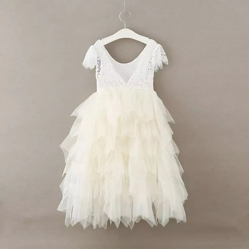 A white toddler girl's party dress with a lace V-neck bodice, ruffled cap sleeves, and a tiered tulle skirt, hanging on a white wooden hanger against a solid taupe background.