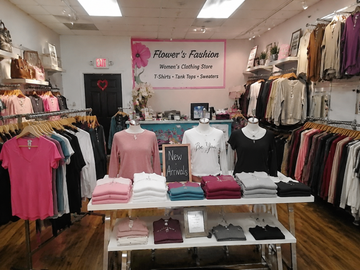 A retail clothing store interior with racks of garments on both sides and a central display table with folded shirts and mannequins wearing tops. A pink sign on the back wall reads 'Flower's Fashion Women's Clothing Store' with 'New Arrivals' on a small chalkboard.