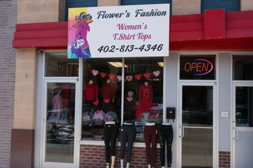 Front exterior of a clothing store named Flower's Fashion, featuring a large white sign with the store name, phone number, and a colorful fashion illustration. The storefront has glass windows displaying women's tops and leggings on mannequins, a red awning, and an illuminated neon 'OPEN' sign.